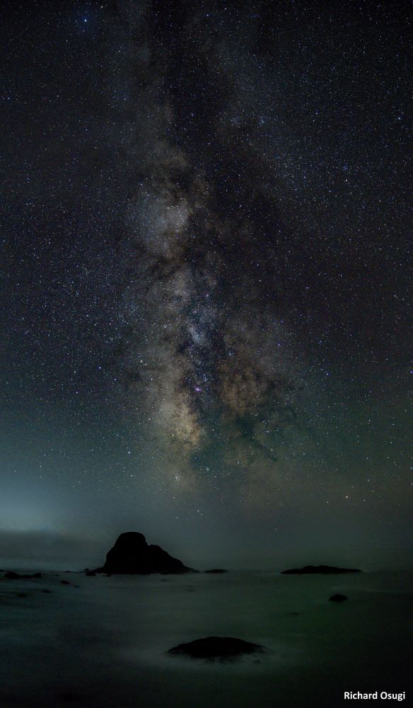 A night sky with the milky way glows above large rocks and the ocean.